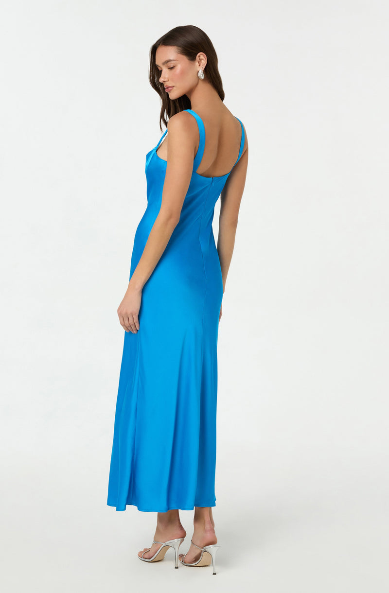 A woman is wearing a long, bright blue dress with thin shoulder straps, fitted at the waist and flowing down to the floor. The dress has a slightly shiny texture and a low, open back. She is also wearing large, elegant earrings and high-heeled, open-toed shoes in a light color.