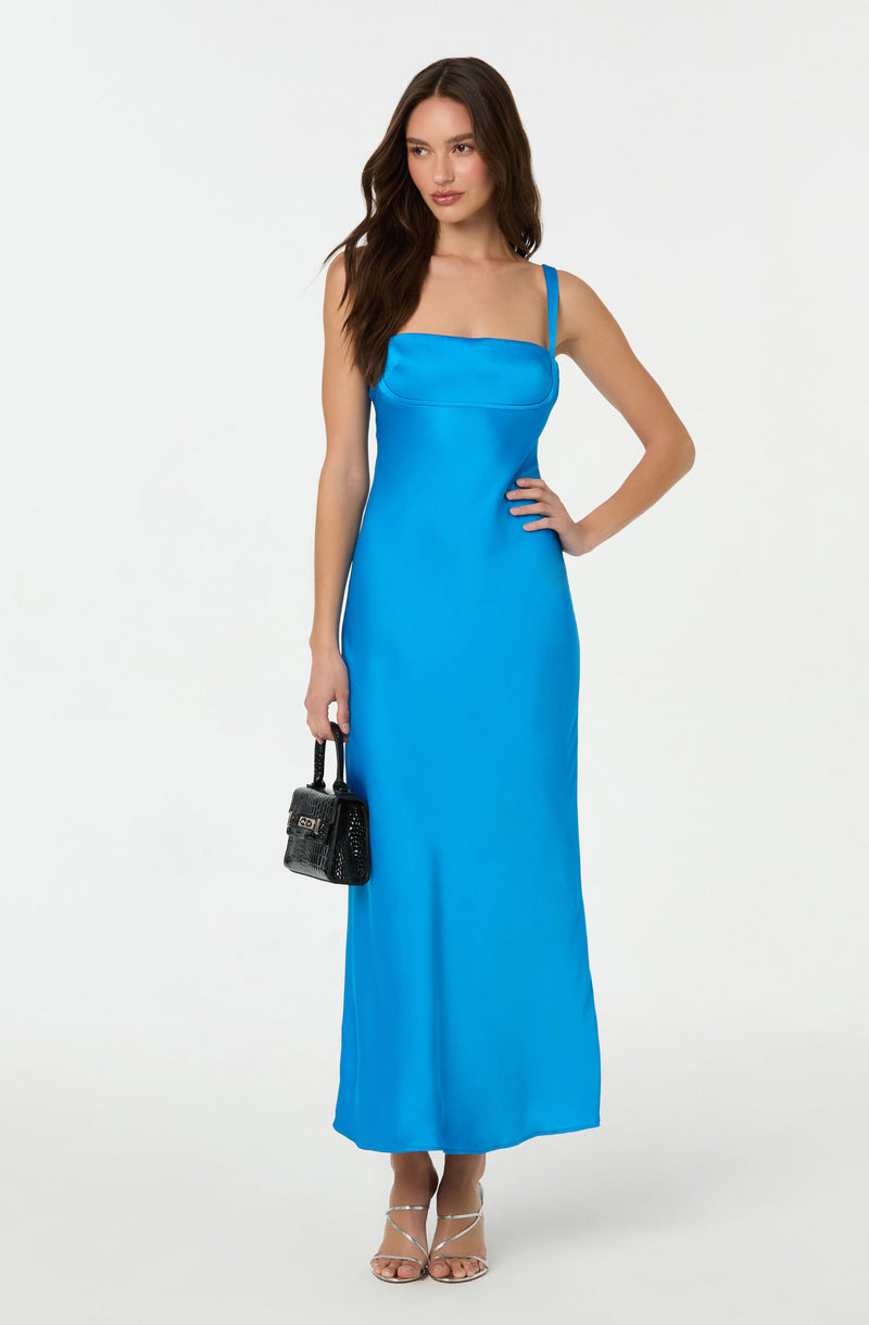 A woman is wearing a long, bright blue sleeveless dress with spaghetti straps and a fitted bodice. She is also holding a small black handbag with a top handle and wearing silver strappy high-heeled sandals.