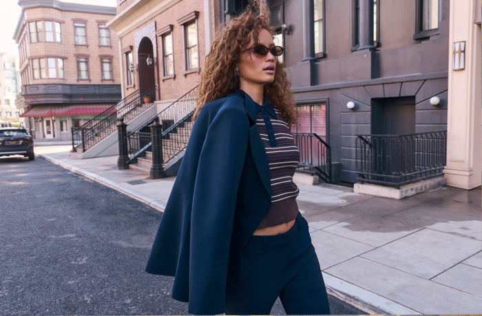 Woman in striped crop top, navy blazer draped over shoulders, brown pants, sunglasses.
