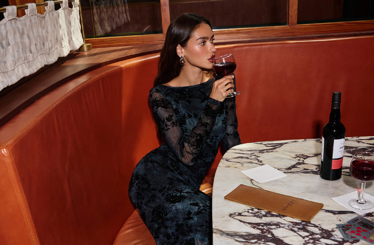 Woman in a black lace long-sleeve dress drinking red wine at a marble table.