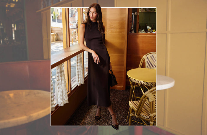 A woman wearing a sleeveless, high-neck, dark brown midi dress with a satin-like sheen, standing in a cafe setting with wooden tables and chairs.