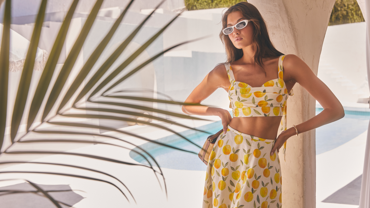 Model wearing the Myria Floral Tie-Strap Cropped Bustier Top and Myria Floral Linen A-Line Midi Skirt in a yellow lemon print two-piece set, styled poolside in a Mediterranean setting — vacation outfit ideas from ASTR the Label.