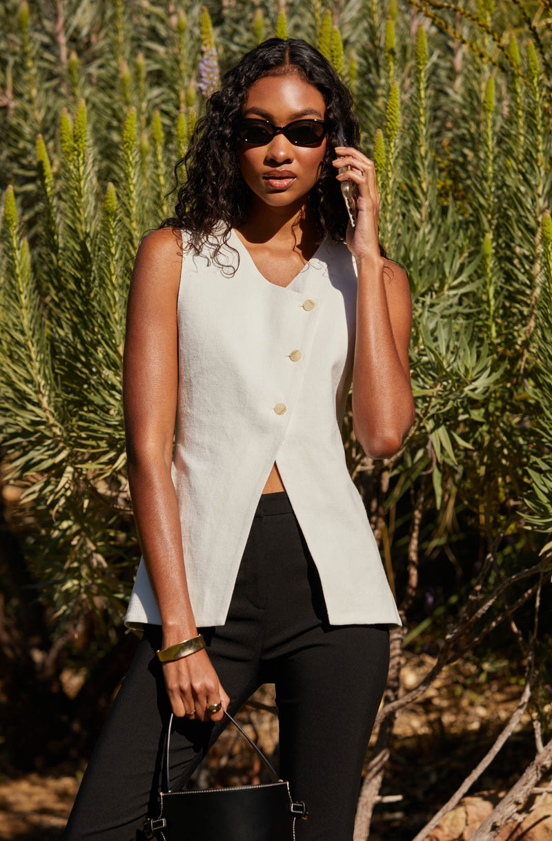 A woman is wearing a sleeveless, off-white, button-front top with a high neckline, paired with black pants. She has on dark sunglasses and is accessorizing with a gold bracelet and ring, and is holding a black handbag. She is standing outdoors in front of green, spiky plants.