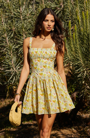 A woman is wearing a sleeveless, fitted dress with a square neckline and wide shoulder straps. The dress has a floral pattern with light colors, primarily yellow, green, and white, and features a gathered, skirted bottom that flares out from the waist. She is accessorizing with large, round earrings, a bracelet, and is holding a woven straw handbag.