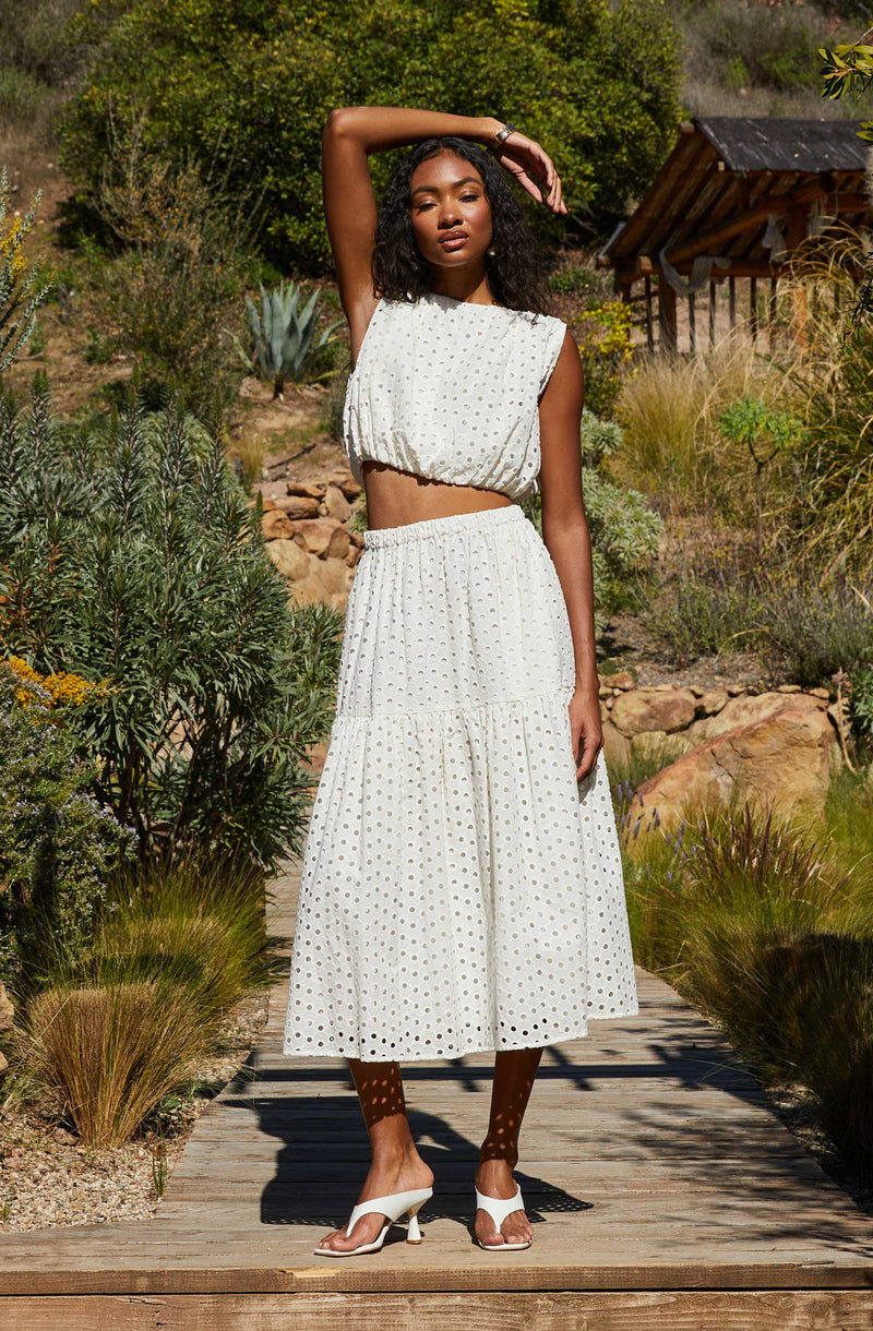 A woman is wearing a white two-piece outfit with a sleeveless, cropped top and a high-waisted, ankle-length skirt. The fabric of both pieces features a subtle dotted pattern. She is also wearing white, open-toe heels.