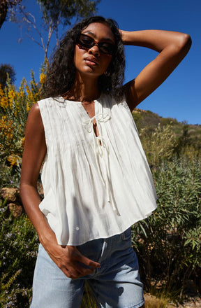 A woman is wearing a sleeveless, white, lightweight top with vertical pleats and a tie-front neckline. She pairs it with light blue denim shorts and accessorizes with dark sunglasses.