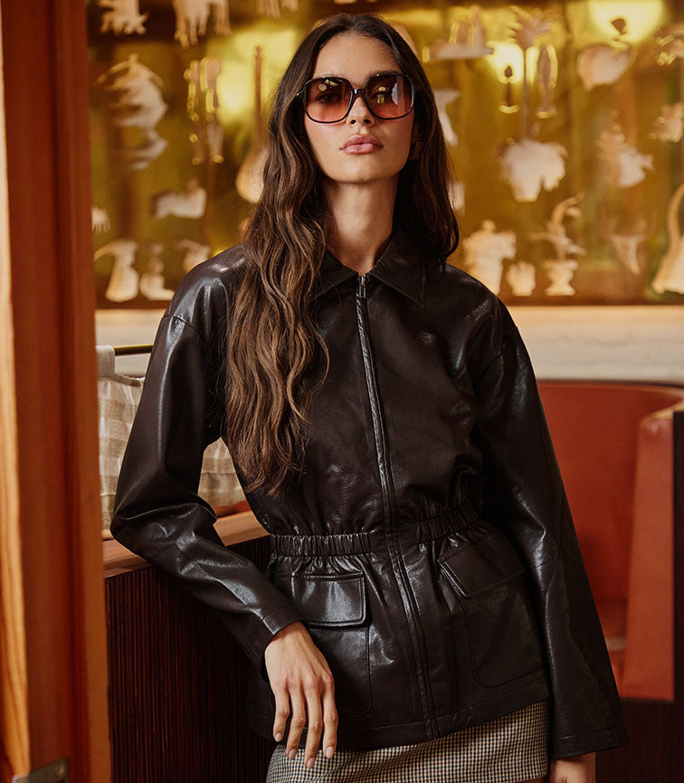 Woman in a black leather jacket with a cinched waist, wearing large sunglasses.