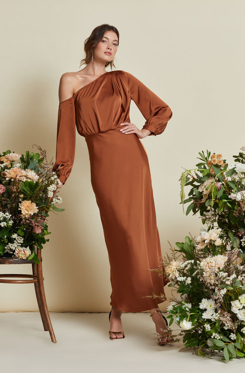 A woman is wearing an elegant, floor-length, satin dress in a warm copper-brown color. The dress features an asymmetrical design with one shoulder, long puffed sleeves gathered at the wrists, and a fitted waist. She is standing beside floral arrangements, dressed in high-heeled sandals, and has her hair styled in loose waves.