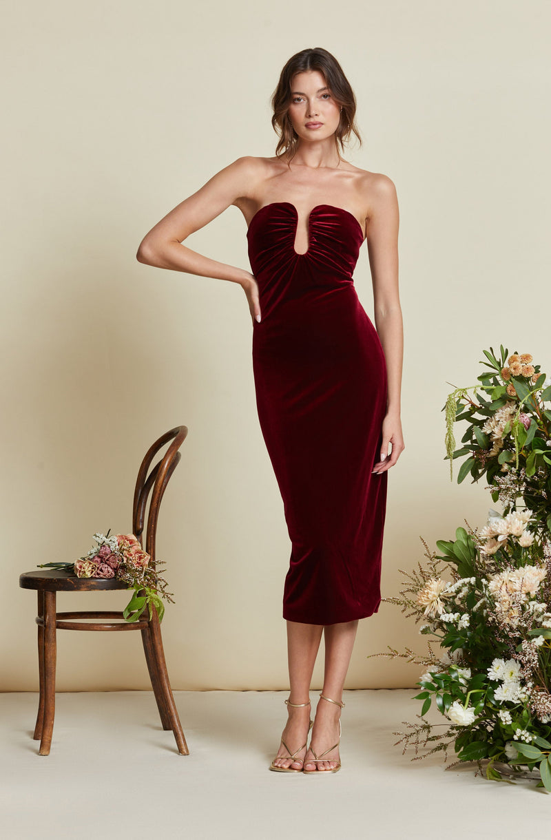 A woman is wearing a strapless, deep red velvet midi dress with a sweetheart neckline that features a central slit detail. The dress is fitted and has a gathered, ruched design at the bust. She pairs it with delicate, open-toe high-heeled sandals.