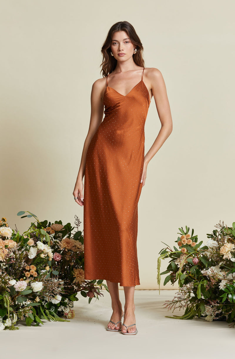 A woman is wearing a rust-colored, satin slip dress with thin spaghetti straps. The dress features a subtle dotted pattern throughout and has a midi length. She is standing among floral arrangements with her right hand resting by her side and her left hand in her pocket.