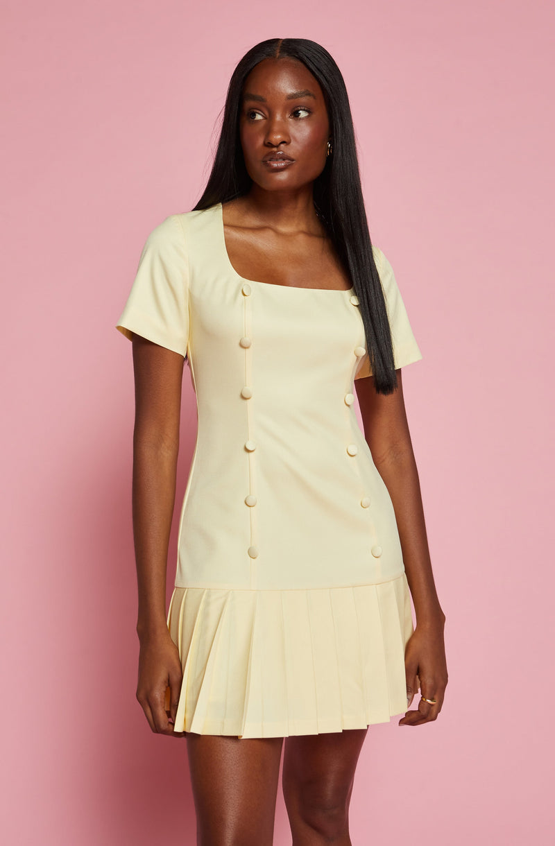 A woman is wearing a light yellow dress with short sleeves. The dress features decorative buttons along the front, a square neckline, and a pleated hem that adds a structured look to the bottom of the dress.