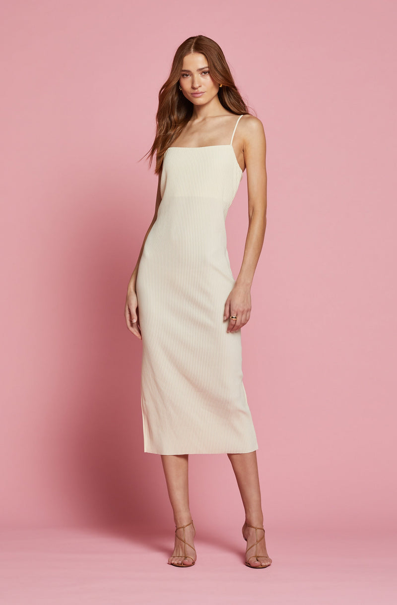 A woman is wearing a sleeveless, ankle-length dress with thin spaghetti straps. The dress is light-colored, possibly cream or off-white, with a ribbed texture. She is also wearing strappy, high-heeled sandals that are nude in color.