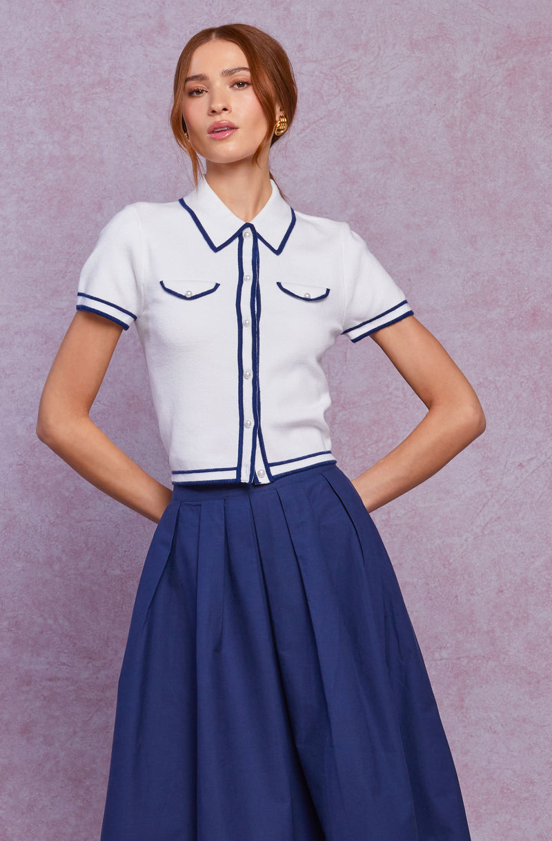 A woman is wearing a white short-sleeved shirt with navy blue trim along the collar, button placket, sleeve cuffs, and two chest pockets. She is also wearing a high-waisted, wide-leg navy blue skirt with pleats.