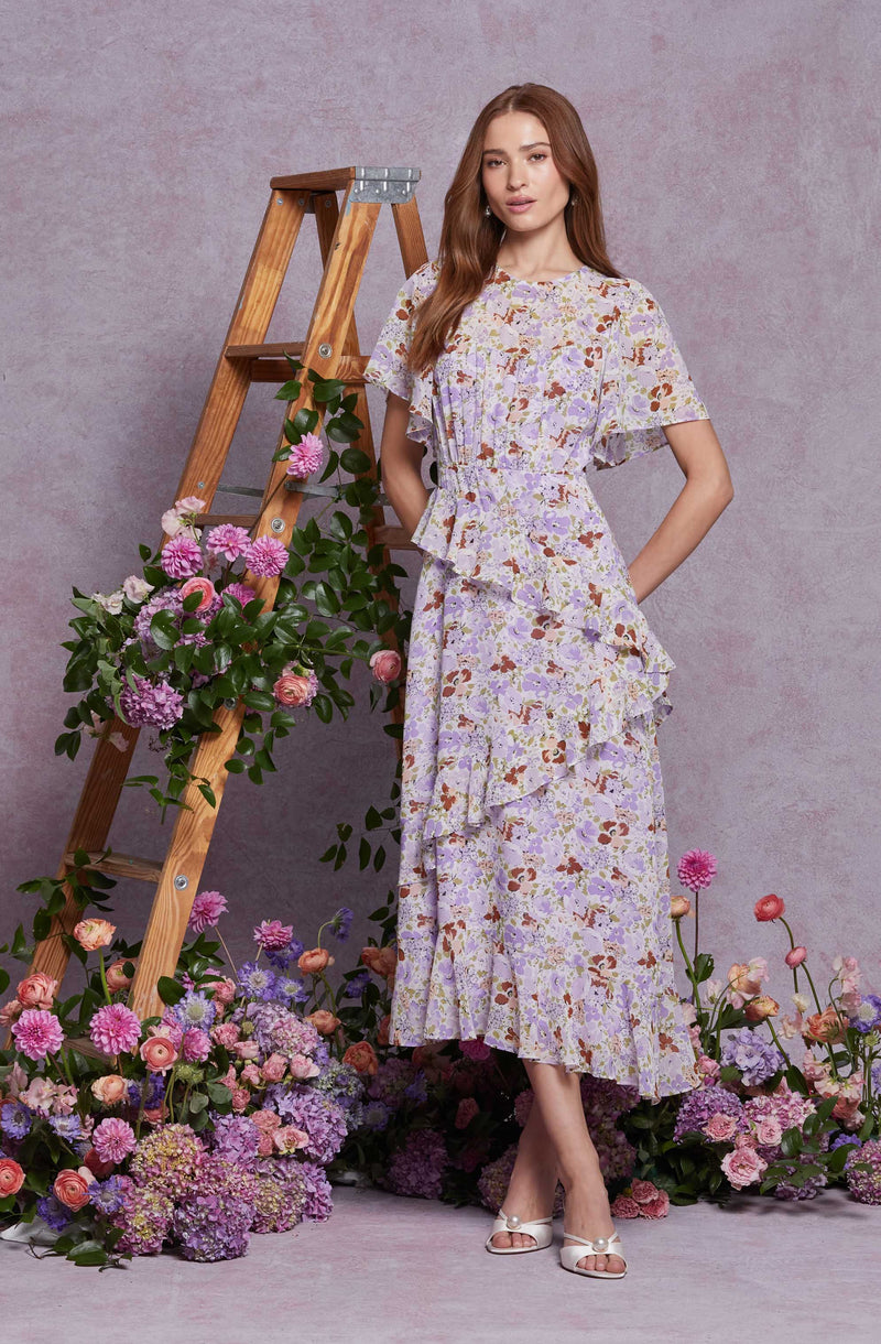 A woman is wearing a long, flowing dress with a floral pattern featuring shades of purple, pink, and brown. The dress has ruffles along the front, waist, and hemline, creating a layered effect. She is also wearing white open-toe shoes with a bow detail. The background is a soft pink, and she stands in front of a wooden ladder decorated with pink and purple flowers, with more flowers scattered on the floor around her.