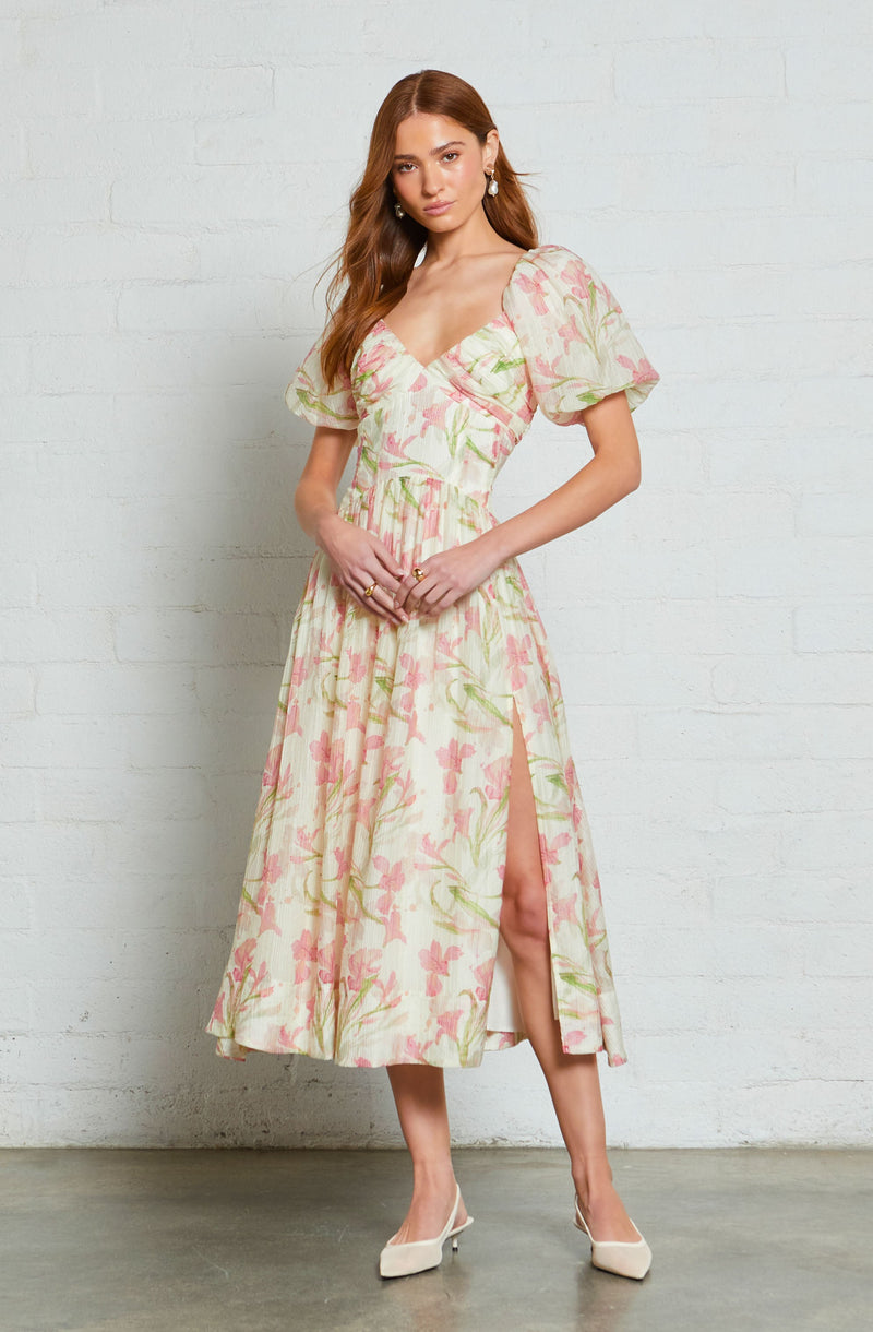 A woman is wearing a midi dress with a floral pattern in light pink, green, and cream colors. The dress features a fitted bodice with a sweetheart neckline, short puffed sleeves, and a high slit on the side revealing part of her leg. She is also wearing white heels and earrings.