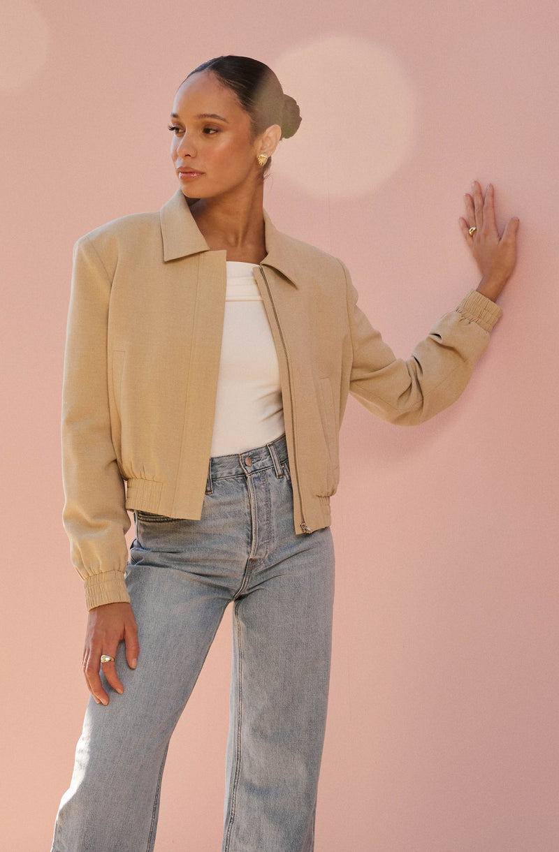 A woman is wearing a beige bomber jacket with ribbed cuffs and waistband, a white top underneath, and light blue high-waisted jeans. She has her hair styled in a bun and is standing against a pink background, with her right hand resting on the wall.