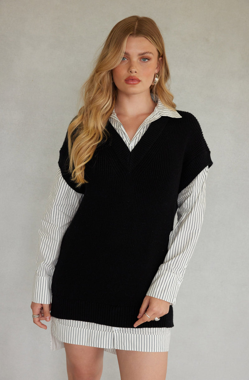 A woman is wearing a white button-up shirt with thin black vertical stripes, layered under a black sleeveless knit sweater vest. The shirt's collar and the bottom hem are visible, with the shirt extending below the vest. The woman has long, wavy blonde hair and is posing against a plain light gray background.