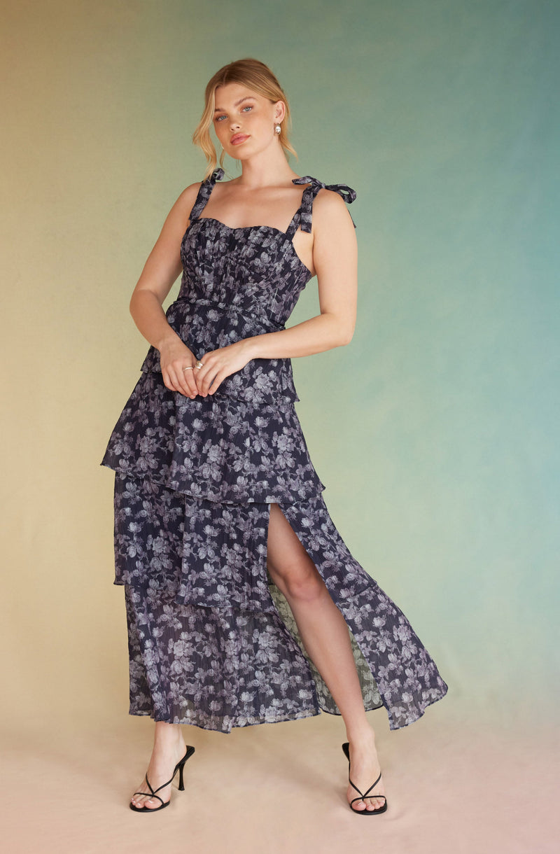 A woman is wearing a sleeveless dress with a dark background and a light floral pattern. The dress has shoulder straps tied into bows and features a tiered skirt with a high slit, revealing her leg. She is accessorized with earrings and rings, and is wearing black high-heeled sandals.