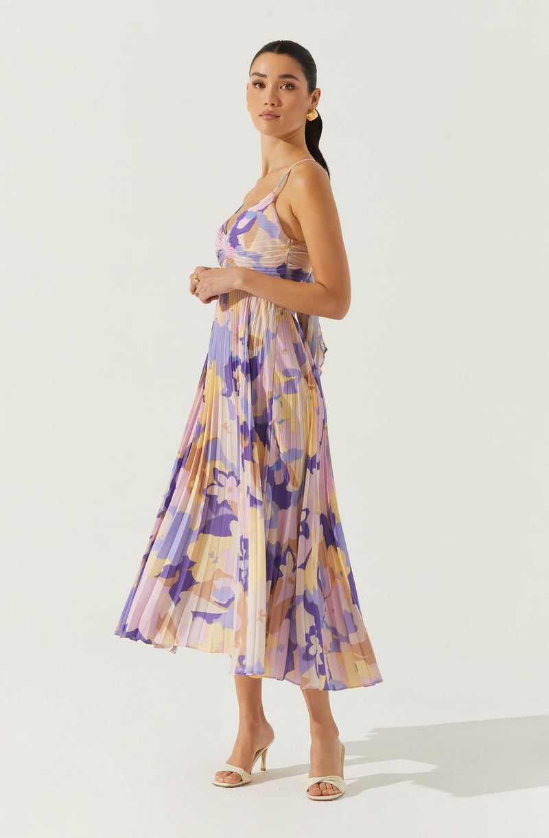 A woman is wearing a sleeveless, spaghetti strap dress with a colorful, abstract floral pattern in shades of purple, yellow, pink, and beige. The dress is pleated and flows down to her ankles. She is also wearing beige open-toed high-heeled sandals.