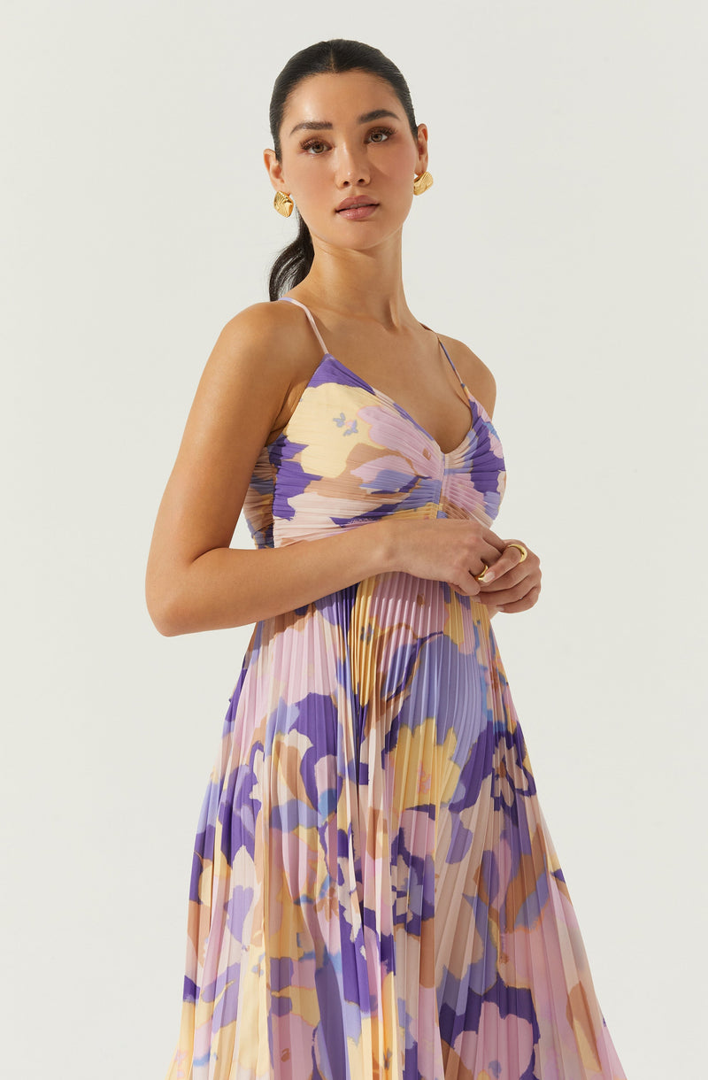 A woman wears a sleeveless, pleated maxi dress with a floral pattern in shades of purple, pink, yellow, and beige. The dress features thin spaghetti straps and a gathered, ruched bodice. She accessorizes with large, gold, textured hoop earrings and a simple gold ring on her left hand.
