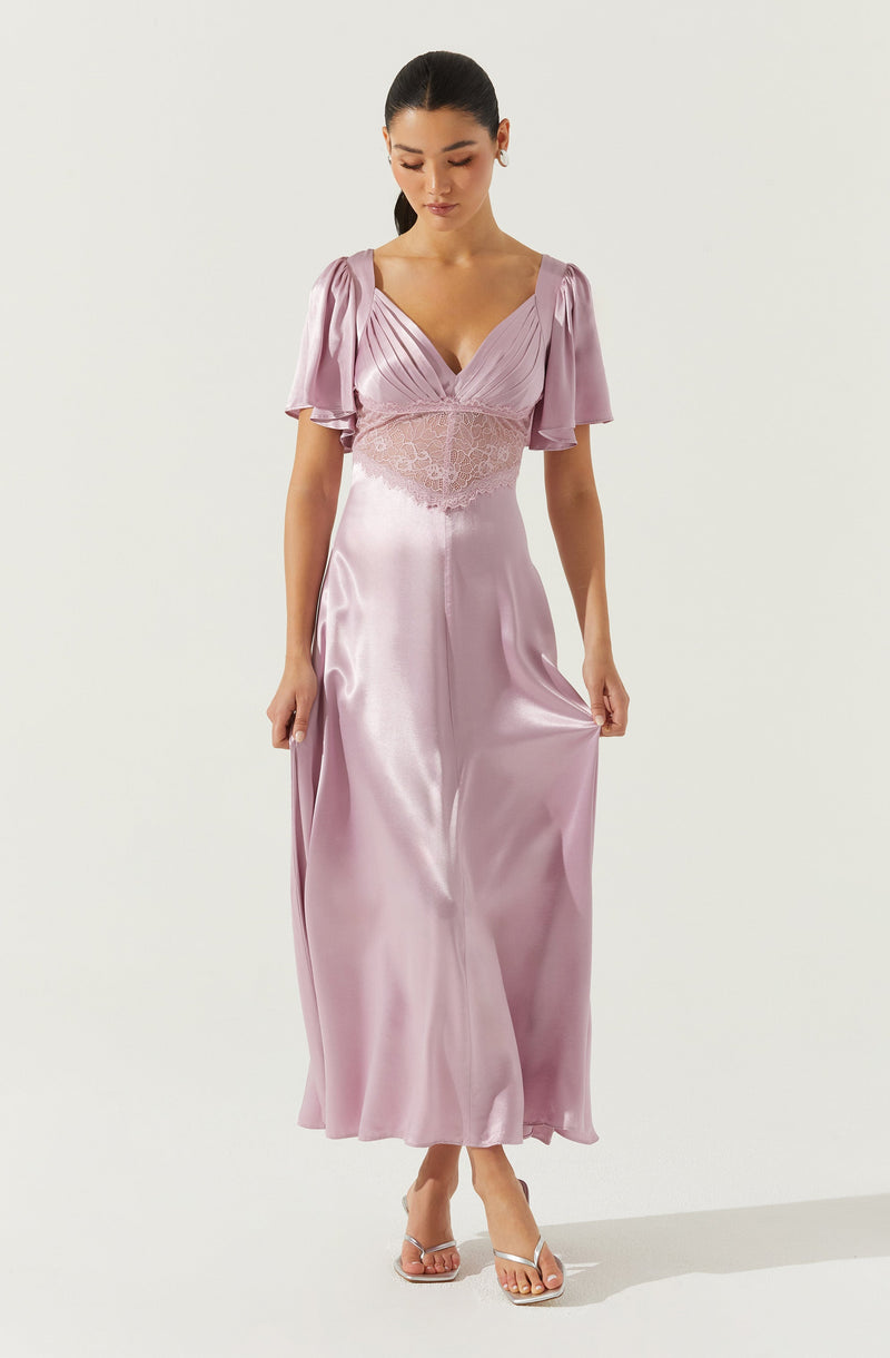 A woman is wearing a long, flowing gown made of silky, shiny fabric in a soft, pastel pink color. The dress features a deep V-neckline with pleated detailing at the bust, short puffed sleeves with a slight sheen, and a lace or mesh panel around the waist area. She is also wearing white high-heeled sandals with thin straps.