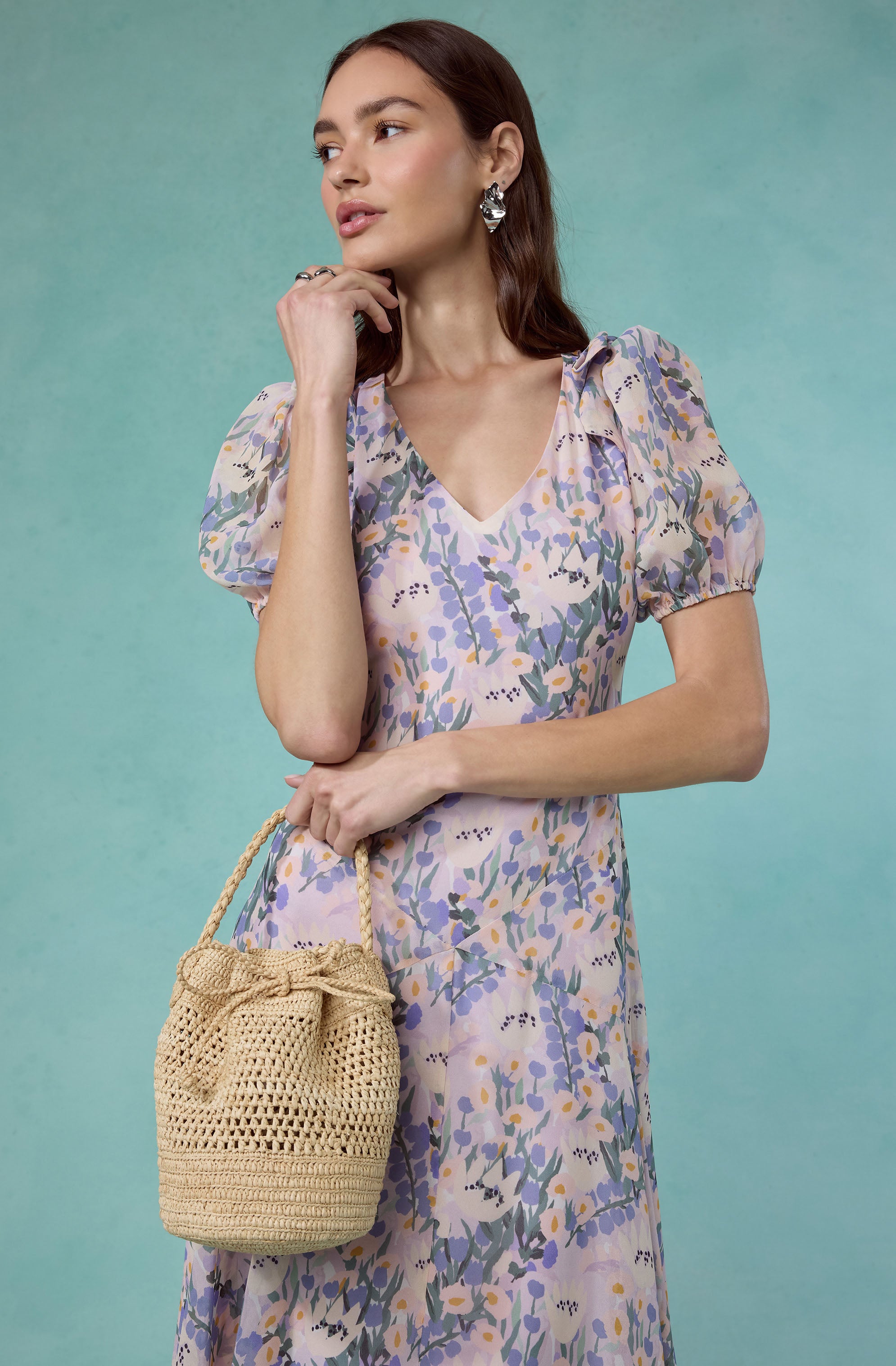 Woman in a pastel purple floral dress with puffed sleeves and a beige woven handbag.