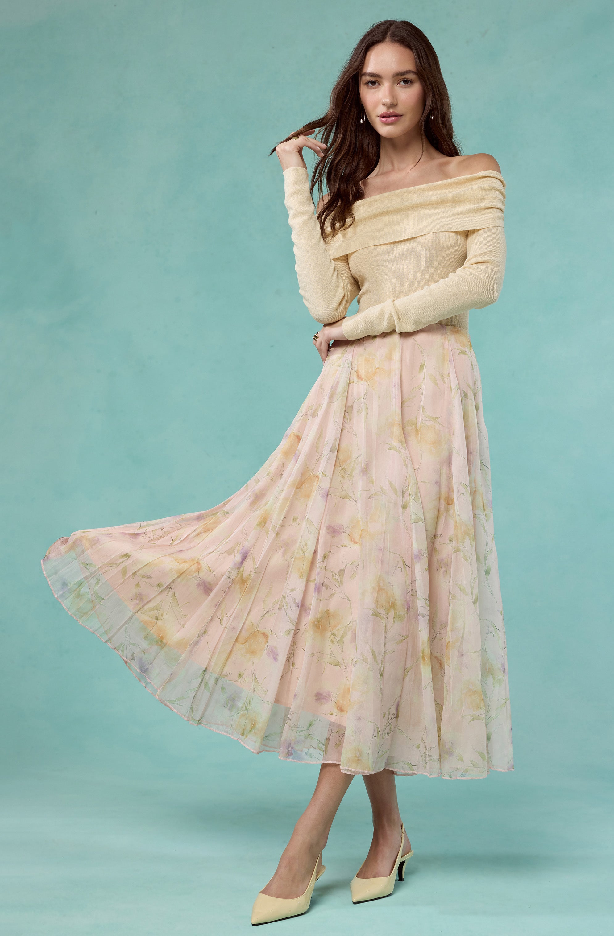 Woman wearing a cream off-shoulder sweater and a pastel floral pleated midi skirt with pale yellow heels.