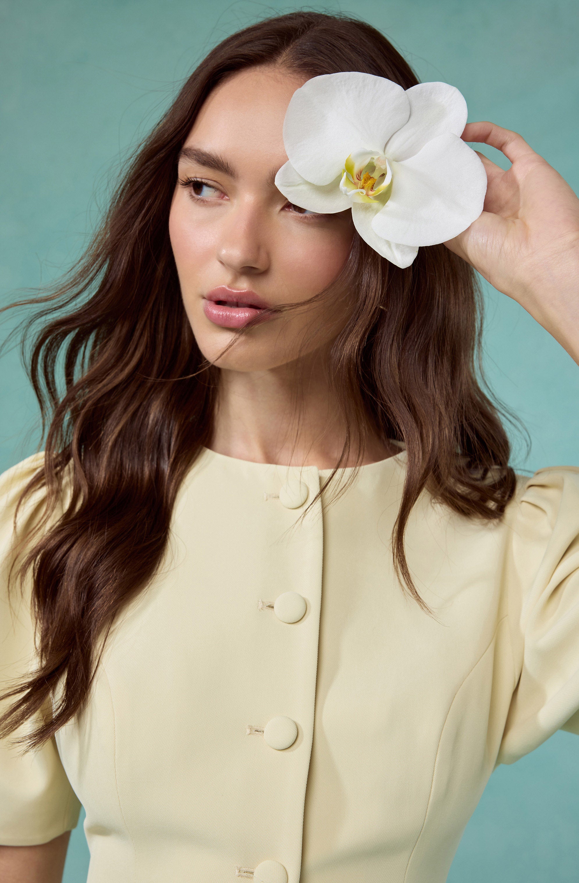 Woman in a cream-colored dress with large buttons, holding a white orchid by her ear.