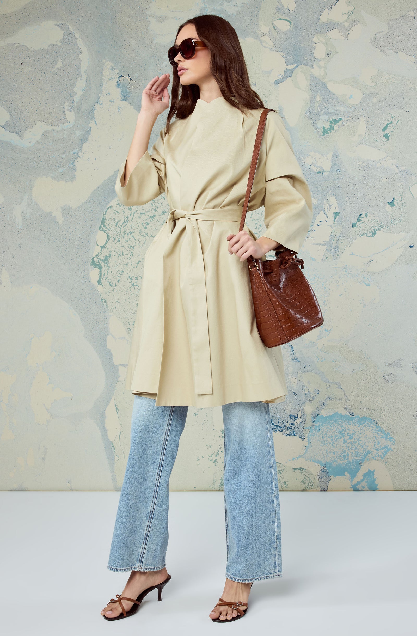 Woman in beige trench coat, light blue jeans, brown sandals, and large sunglasses.