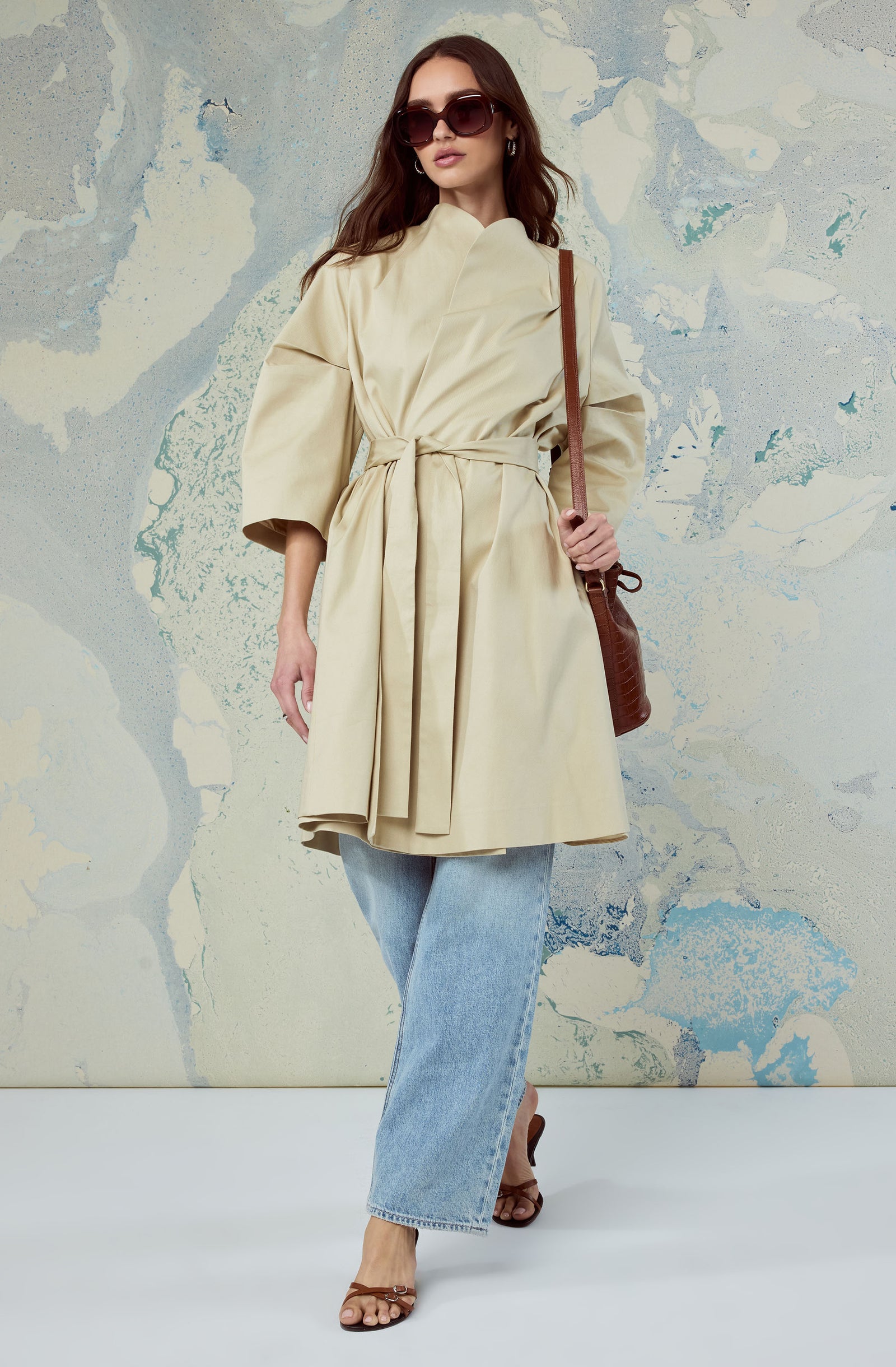 Woman wearing a beige belted trench coat, light blue jeans, and brown sandals, carrying a brown shoulder bag, with dark sunglasses.