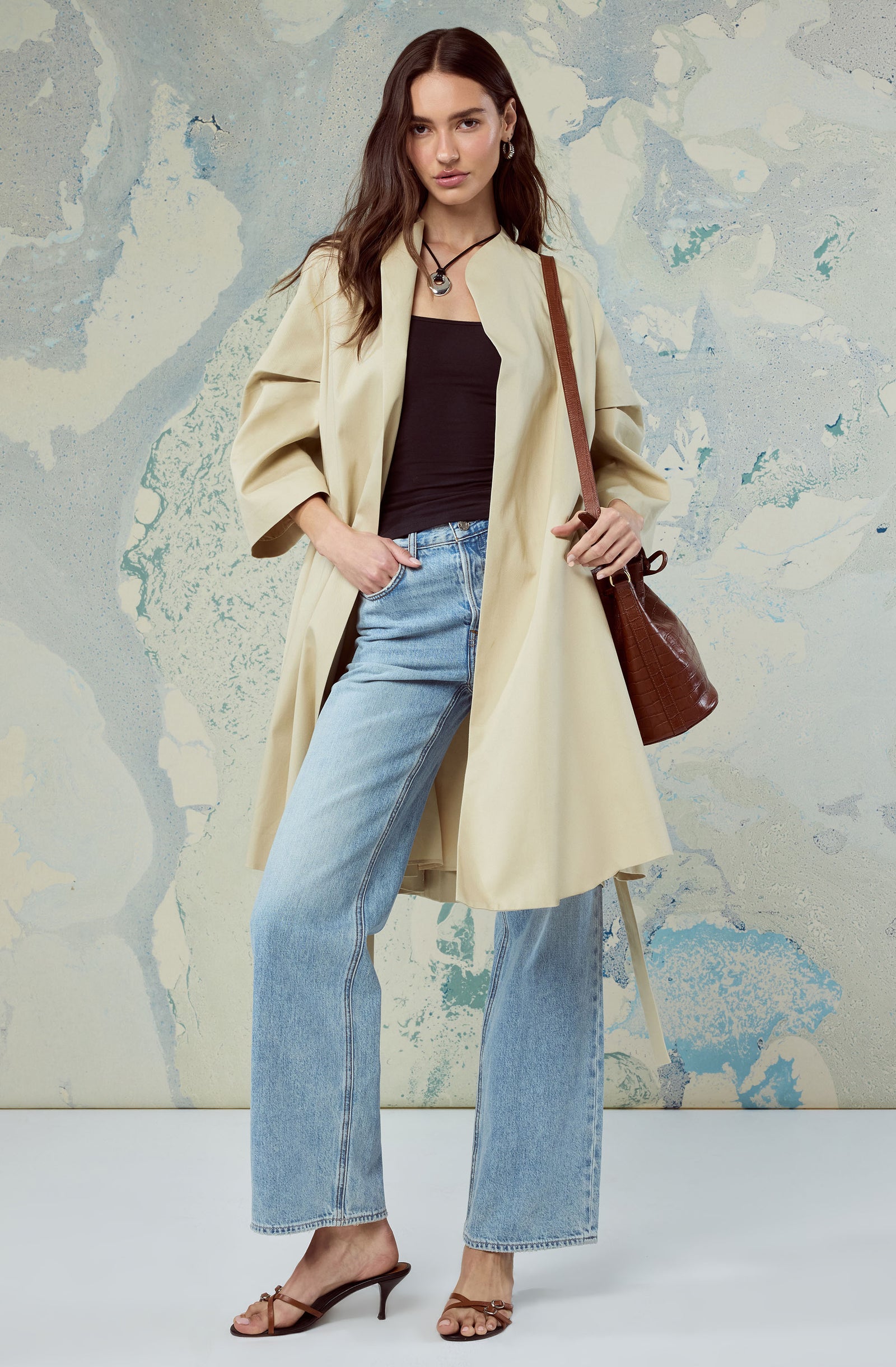 Woman wearing a beige trench coat, black top, light blue jeans, brown heels, and carrying a brown handbag.