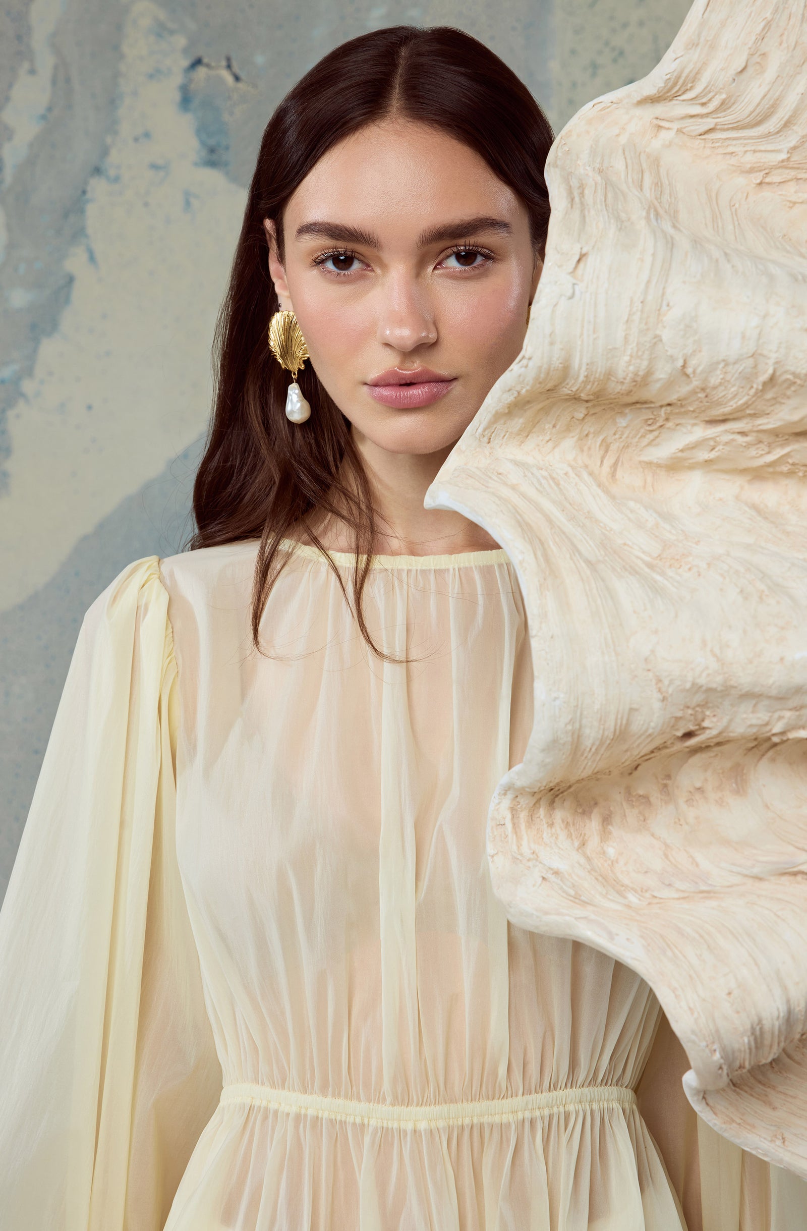 Woman wearing a light cream, flowing dress with pleats and puffed sleeves, large gold and pearl earring.
