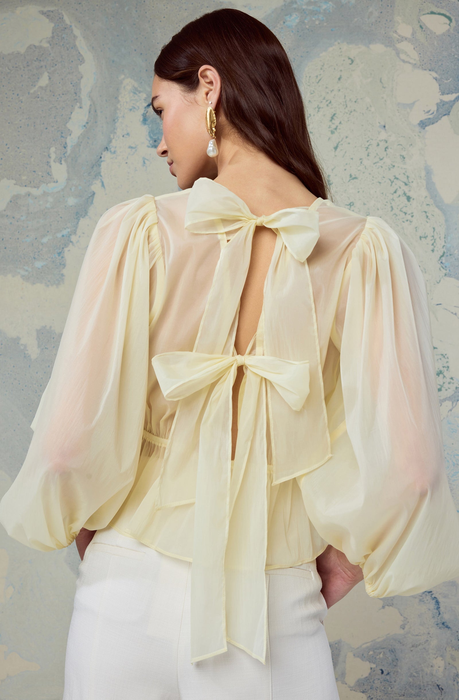 Woman wearing a cream, sheer blouse with puffed sleeves and large bows on the back.