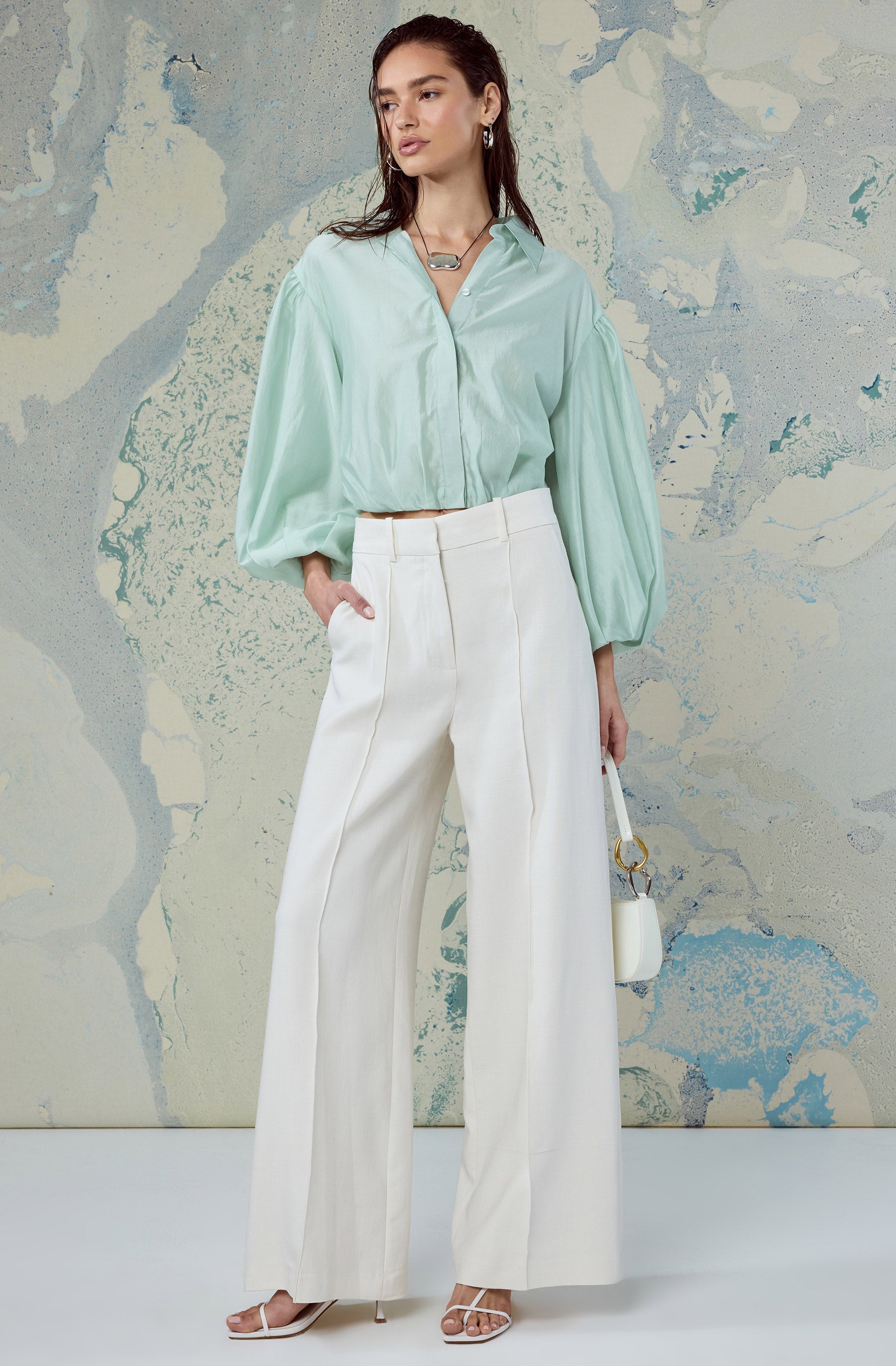 Woman in a pastel green oversized blouse with puffed sleeves, high-waisted white wide-leg trousers, and white heels.