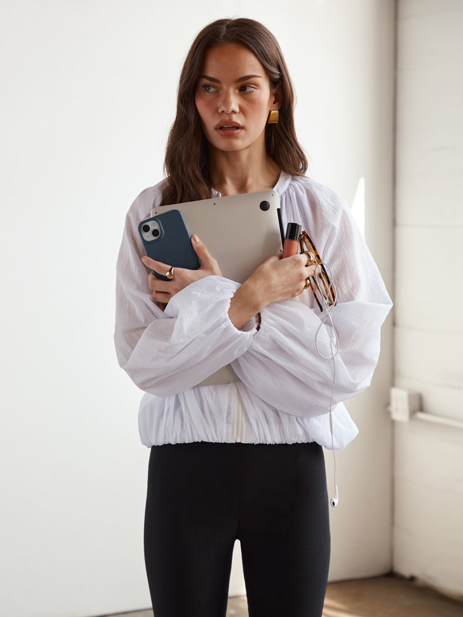 Woman in white cropped jacket holding a tablet, phone, and lip balm.