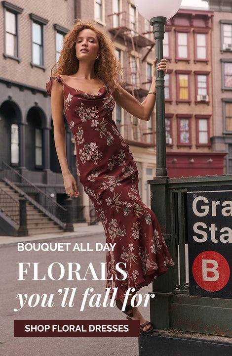 Young woman in a long, burgundy floral dress standing on a city street near a lamp post.