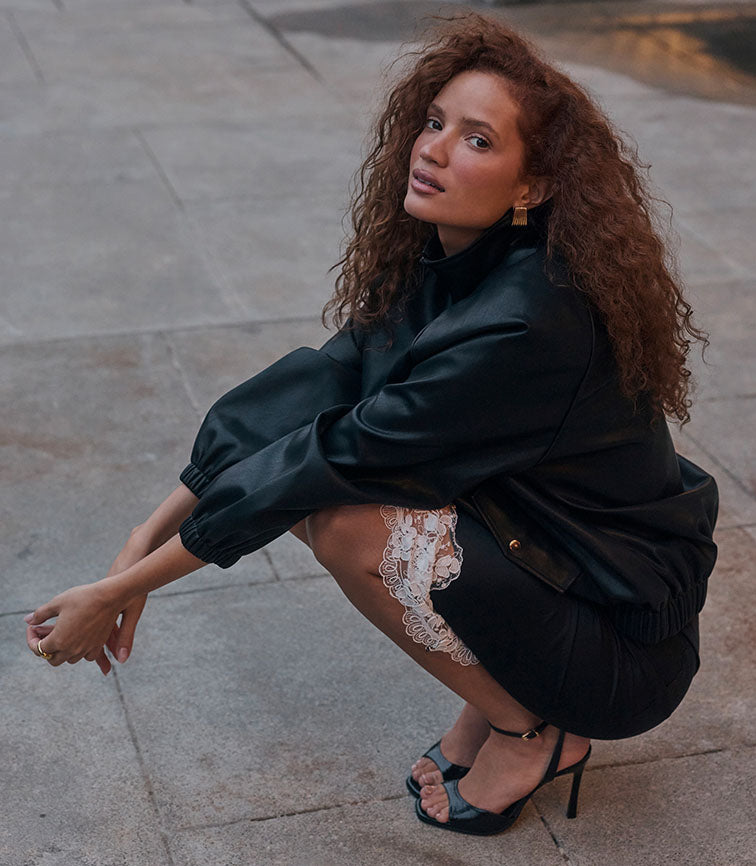 Woman in black leather jacket, lace shorts, and high heels crouching.