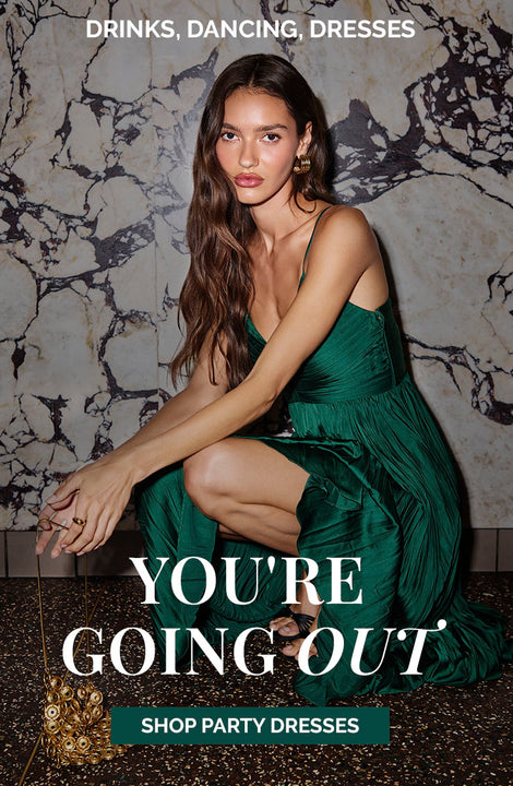 Woman in a shiny emerald green party dress with thin straps, sitting against a marble wall.