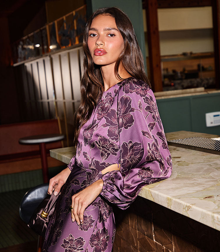Woman in purple floral satin blouse and matching skirt, standing at bar counter.