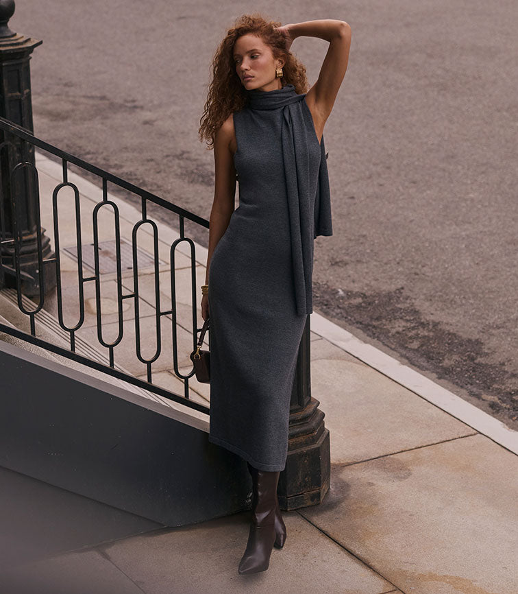 Woman in a sleeveless, high-neck, dark gray dress with a long scarf, paired with dark boots.