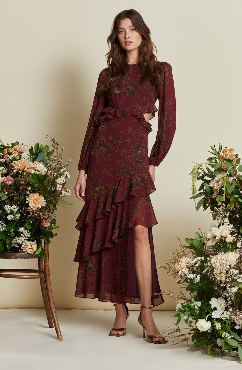 A woman is wearing a long, flowy, burgundy dress with a floral pattern. The dress has long sleeves and a ruffled hem that cascades asymmetrically down the front, with a high slit revealing her leg. She is also wearing nude-colored high-heeled sandals.
