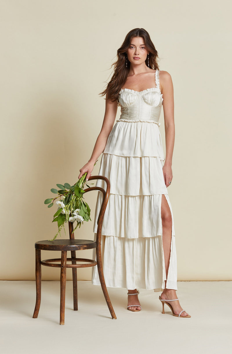 A woman is wearing a sleeveless, cream-colored tiered dress with ruffle details on the bodice and shoulder straps. The dress has a high slit on the side, revealing her leg. She is also wearing light-colored, open-toe high heels. The outfit appears to be elegant and summery.