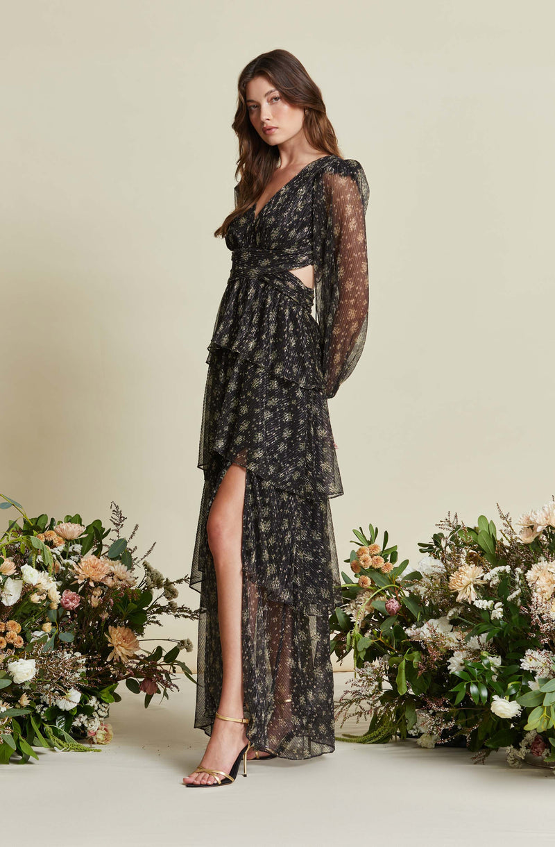 A woman is wearing a long, black, floral dress with multiple layered ruffles. The dress has long, sheer sleeves and features a high slit on one side, revealing her leg. The fabric is semi-transparent, with delicate, small floral patterns throughout. She is also wearing high-heeled, open-toe sandals with ankle straps. The background is a light, neutral color, and there are floral arrangements on both sides of her.