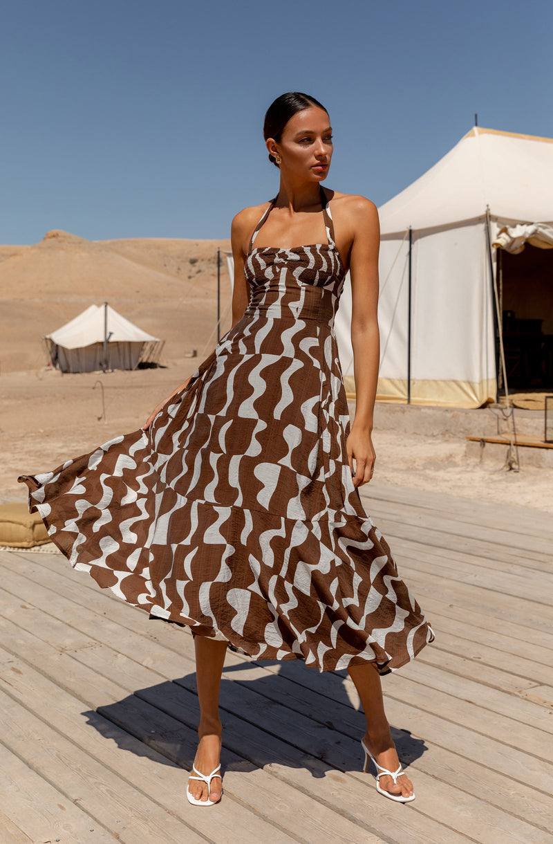A woman is wearing a sleeveless, halter-neck dress with a brown and white geometric pattern. The dress features a fitted bodice and a flowing, asymmetrical hemline that reaches mid-calf length. She is also wearing white strappy high-heeled sandals.