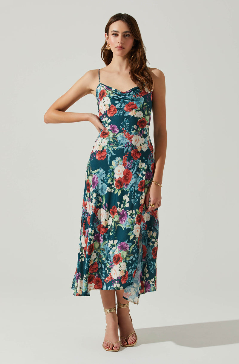 A woman is wearing a sleeveless, ankle-length dress with a teal background and a vibrant floral pattern consisting of red, white, purple, and blue flowers. The dress features thin spaghetti straps and a slightly ruched neckline. She pairs the dress with beige high-heeled sandals and gold jewelry, including earrings, a bracelet, and a ring.