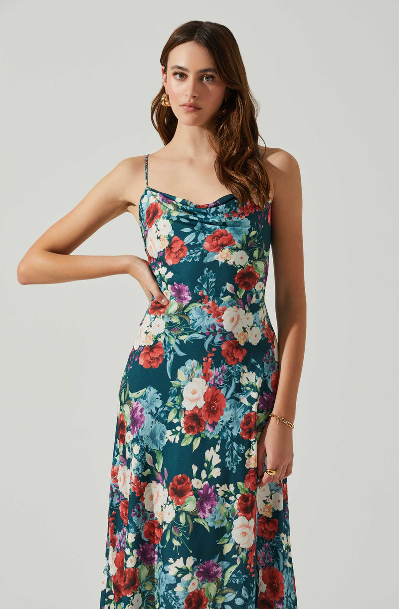 A woman is wearing a sleeveless, floor-length dress with thin spaghetti straps. The dress features a vibrant floral pattern with large red, white, purple, and blue flowers set against a dark teal background.