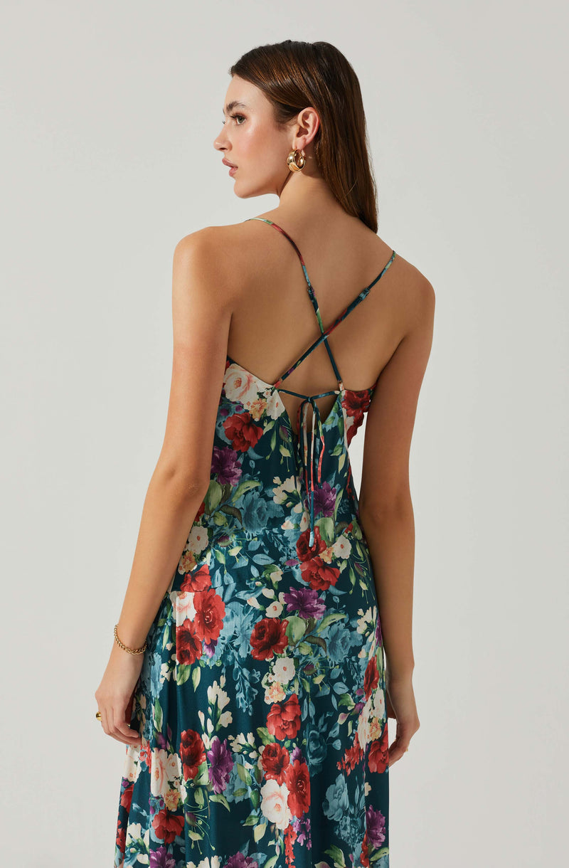 A woman wearing a sleeveless, floral-patterned dress with spaghetti straps. The dress features a dark background with vibrant red, purple, white, and green floral prints. The back of the dress has thin, crisscrossing straps and an open back design with a tied bow at the center. She has a gold bracelet on her left wrist and large gold hoop earrings.