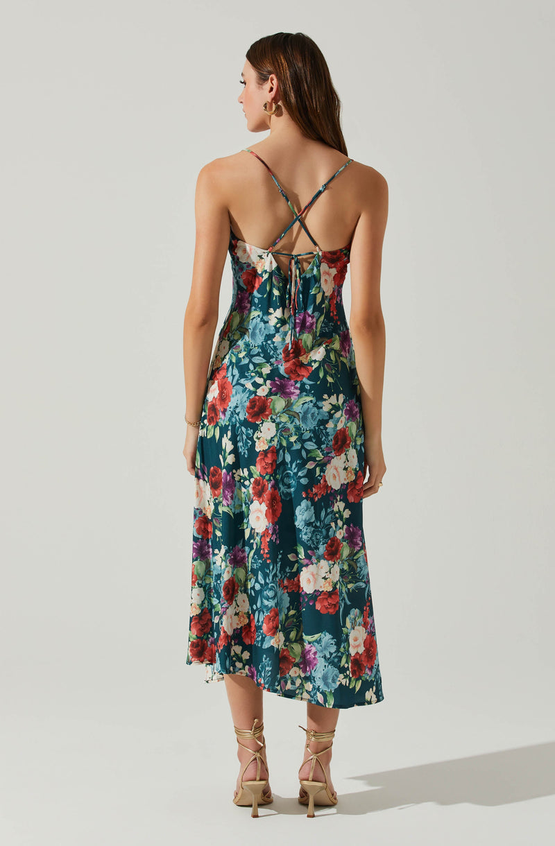 A woman is wearing a sleeveless, ankle-length dress with a vibrant floral pattern in red, white, purple, and green on a dark blue background. The dress features thin, adjustable spaghetti straps that cross at the back and tie into a bow. The fabric appears lightweight and flowy. She is also wearing nude-colored, lace-up high-heeled sandals with thin straps wrapping around her ankles.
