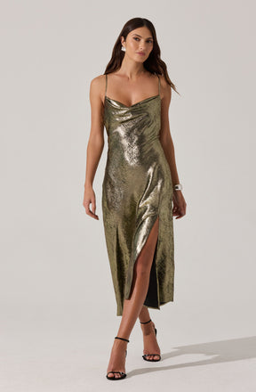 A woman is wearing a long, gold metallic dress with thin shoulder straps. The dress features a cowl neckline and a high slit on the front left side. She has paired the outfit with black high-heeled sandals.
