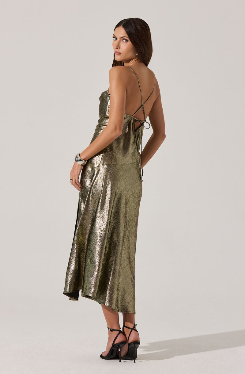 A woman is wearing a long, metallic, gold-colored dress with a shiny, textured fabric. The dress features thin spaghetti straps and an open back with a lace-up detail. She is also wearing black high-heeled sandals with ankle straps.